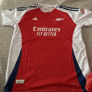 Arsenal hemmatröja Adidas strl 32 - Snygg Arsenal hemmatröja från Adidas med klassisk röd front, vita ärmar och blå detaljer. Tröjan har korta ärmar, broderat klubbmärke och Adidas-logga, samt Premier League-märke på ärmen. Materialet är lätt och andas, perfekt för match eller träning.