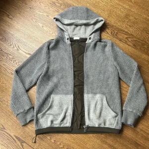 Moncler cardigan  - Snygg grå hoodiejacka från Moncler med quiltad mörkgrön baksida och insida. Jackan har stickade ärmar och huva i grov struktur, stora fickor fram och dragkedja. Perfekt för dig som gillar streetstyle och vill ha något unikt och bekvämt. Skick 8/10