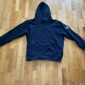 Svart hoodie från Stone Island - Svart hoodie från Stone Island med klassisk patch på vänster ärm. Tröjan har huva med snörning, stor magficka och långa ärmar. Tillverkad i mjuk bomull, perfekt för en chill och stilren look. Det är Medium men sitter som S