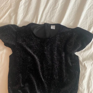 Svart glittrig blus - Supersnygg svart blus som är glittrig. Blusen har korta ärmar som är volangiga och rund halsringning, perfekt på fester. Materialet är mjukt och skimrande, vilket ger en lyxig känsla. Aldrig andvänt. Pris kan diskuteras 