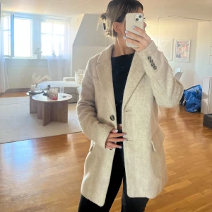 Beige kappa från H&M - Snygg beige kappa från H&M i storlek XS. Klassisk modell med krage, knäppning framtill och fickor på sidorna. Jackan har en mjuk ullkänsla och är perfekt för dig som gillar stilrena och tidlösa plagg.