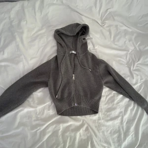 Grå croppad hoodie från Zara - Grå croppad hoodie från Zara med dragkedja framtill och snörning i huvan. Tröjan har ribbade, långärmade ärmar och är stickad i mjukt material.