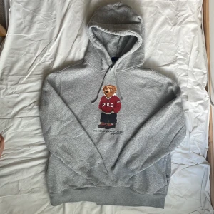 Grå Polo Bear hoodie från Ralph Lauren - Säljer en grå hoodie från Ralph Lauren med ikoniska Polo Bear-trycket på bröstet. Tröjan har huva med snörning och är tillverkad i mjuk bomull. Perfekt för dig som gillar streetwear och vill ha en chill look med en klassisk touch. ⚠️ Obs!!! Tröjan har en blekningsfläck under trycket, se bild 5. Resten av tröjan är i väldigt bra skick