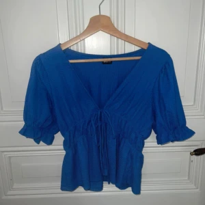 Blå blus med knyt från Gina Tricot - Supersöt blå blus från Gina Tricot med v-ringning och knyt framtill. Modellen har korta puffärmar med volangkant och en lätt strukturerad yta. Perfekt för dig som gillar färg och vill ha en feminin touch på outfiten.