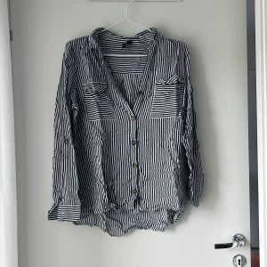 Randig skjorta från Vero Moda L - Snygg skjorta från Vero Moda i svart och vitt med smala ränder. Skjortan har knappar framtill, två bröstfickor och lång ärm som kan vikas upp. Tillverkad i tunn och luftig bomull, perfekt för en chill och avslappnad stil.