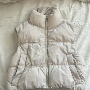 Vit dunväst från H&M Divided XXS - Säljer en fluffig och stilren vit dunväst från H&M Divided i storlek XXS. Västen har hög krage, två fickor framtill och stängs med dragkedja. Perfekt att slänga över en hoodie för en cool lager-på-lager-look.