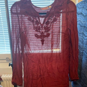 Vinröd blus med broderi från Indiska - Vinröd transparent blus från Indiska med långa ärmar och v-ringning. Blusen har vackra broderade detaljer framtill och är tillverkad i ett lätt och luftigt material. Perfekt för dig som gillar bohemisk och romantisk stil.