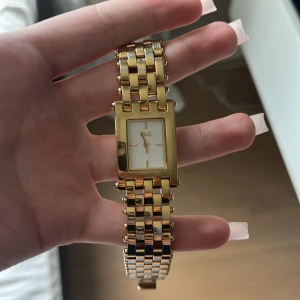 Guldig klocka dolce & Gabbana  - Stilren klocka från D&G med rektangulärt ur i guldton och vit urtavla. Länken är bred och glänsande i metall, vilket ger en lyxig känsla. Minimalistisk design med guldiga visare och markeringar. Perfekt accessoar för att lyfta din outfit. Guldet har tappats lite men det är inget som syns när den bärs.