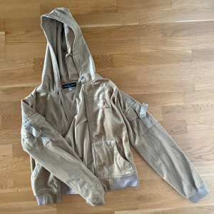 Beige velour hoodie från Baby Phat - Säljer en guldbrun aktig kofta i sammet från Baby Phat. Tröjan har dragkedja, huva med snörning, broderad logga på bröstet och coola fickor med banddetaljer på ärmarna. Perfekt för dig som gillar 00-talsvibe. Passar även S🤎