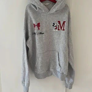 Grå Disney hoodie från H&M - Grå hoodie från H&M med Disney-tryck framtill, featuring Mickey Mouse, röd rosett och stort M. Klassisk känguruficka, huva och mjukt material. Perfekt för dig som gillar en chill och avslappnad stil.