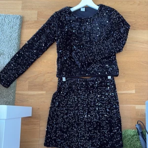 Svart paljettkjol och svart paljetttröja från Lindex - Glittrig svart kortkjol från Lindex, täckt med svarta paljetter som ger en riktigt festlig vibe. Tillsammans med en glittrig svart paljetteöja. Kjolen har rak passform och resår i midjan för extra komfort. Tröjan är i ett skönt material på insidan. Perfekt för dig som vill sticka ut och glänsa på nästa event!