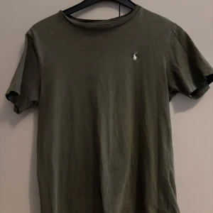 Olivgrön t-shirt från Ralph Lauren - Snygg olivgrön t-shirt från Ralph Lauren med klassisk passform och liten vit broderad logga på bröstet. Tillverkad i mjuk bomull och har rund halsringning samt korta ärmar. Perfekt för en clean och avslappnad stil.