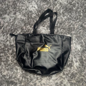 Svart bag från Puma - Stor svart bag från Puma i syntetmaterial med guldig logga framtill. 