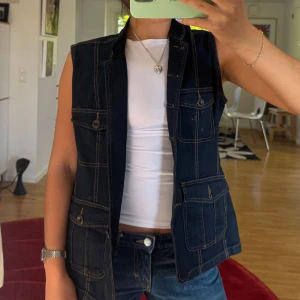 Mörk jeansväst med fickor - Snygg mörkblå jeansväst med kontrastsömmar och fyra stora fickor framtill, alla med knappstängning