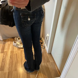 True Religion mörkblå bootcut jeans - Säljer ett par mörkblå jeans från True Religion med ikoniska kontrastsömmar och stora fickor med lock och silverknappar bak. Modellen har låg midja och bootcut-ben💓