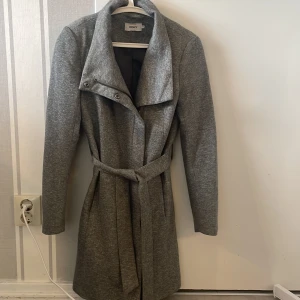 Grå kappa med bälte från ONLY XS - Snygg grå kappa från ONLY i storlek XS. Jackan har en stilren design med bred krage, bälte i midjan och dragkedja samt knappar framtill. Perfekt för dig som gillar en clean look och vill ha något som funkar till många outfits. Nästan oanvänd! Köpt den för 800kr
