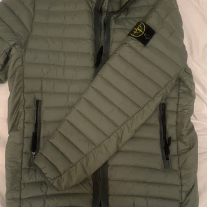 Grön Stone Island Jacka - Säljer en snygg grön dunjacka fràn Stone Island.Jackan har en dragkedja framtill och lánga ärmar.Perfekt för kyligare dagar med sin quiltade design. Den har ocksà praktiska fickor med dragkedja. Den är köpt frän Miinto för 7000. Äkthetscerifikation FINNS! Pris går att diskutera
