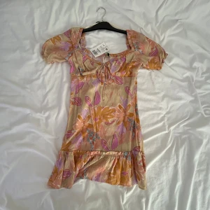 Blommig kortklänning från Positano - Superfin kortklänning från Positano med blommigt mönster i beige, orange och lila toner. Klänningen har puffiga kortärmar, volang nertill och söt knytning vid bysten. Perfekt för dig som gillar färg och romantiska detaljer.