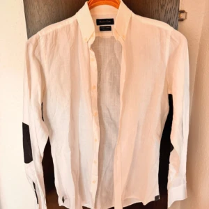 Vit linneskjorta Massimo Dutti - Vit skjorta från Massimo Dutti i svalt linne med klassisk krage och svarta armbågslappar. Långärmad modell med knappar framtill, perfekt för en clean och stilren look. Snygg detalj med kontrast på armbågarna.