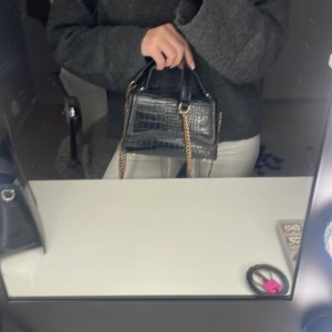 Svart croco handväska med guldkedja - Säljer en svart handväska i croco-mönstrad syntet med guldfärgade kedjedetaljer. Väskan har ett stilrent och kompakt format med dubbla handtag och magnetlås. Perfekt för dig som vill ha en trendig accessoar med lyxig känsla.