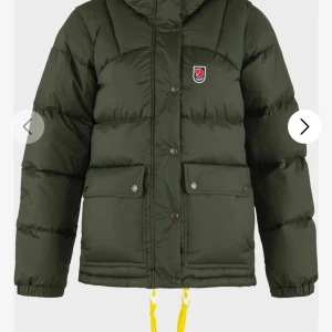 Grön pufferjacka från Fjällräven - Säljer en mörkgrön pufferjacka från Fjällräven med klassisk logga på bröstet. Jackan har stora fickor med knappar, huva och gula snören nedtill. Perfekt för kalla dagar och riktigt skön passform. Fylld med dun och har långa ärmar.
