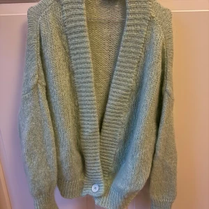 Ljusgrön kofta från American Vintage - Supermjuk och fluffig ljusgrön kofta från American Vintage. Oversized passform med bred ribbad krage, lång ärm och en stor knapp framtill. Stickad i en lyxig mix av mohair, ull och polyamid – perfekt för lager på lager under kyliga dagar.