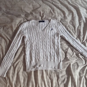 Vit kabelstickad tröja Polo Ralph Lauren - Vit kabelstickad tröja från Polo Ralph Lauren i storlek XS. Tröjan har v-ringning, lång ärm och klassisk blå logga broderad på bröstet. Tillverkad i mjuk bomull och har ribbade muddar vid ärmslut och nederkant. Perfekt för en clean och stilren look.