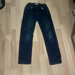 Mörkblå jeansbyxor från Lindex - Snygga mörkblå jeansbyxor från Lindex med klassisk femficksdesign och resår i midjan. Jeansen har raka ben och subtila slitningar framtill för en cool look. Perfekta för dig som gillar enkel och stilren denim.