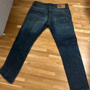 G-Star Jeans - Säljer ett par G-Star Raw 3301 jeans i mörkblå färg med slitningar och ljusa partier på låren. Modellen har rak passform, klassiska fem fickor och knappgylf. Snygga detaljer och robust denimkänsla, perfekta för dig som vill ha en trendig look💯 säljer då de inte passade mig tyvärr. Riktigt snygga byxor i storlek: W36, L32
