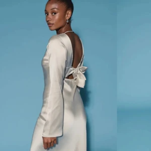 Elegant långklänning i satin med rosett - Supersnygg långklänning i ljus satin med öppen rygg och stor rosett bak. Klänningen har långärmade ärmar och en stilren, figurnära passform. Perfekt för dig som vill sticka ut med en chic och modern look.