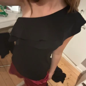 Svart oneshoulder topp från H&M - Snygg svart oneshoulder topp från H&M med volangdetalj längs axeln. Toppen har en asymmetrisk skärning och är tillverkad i ett mjukt, lätt material som sitter bekvämt. Perfekt för dig som vill sticka ut med en stilren och trendig look. Tröjan är i ett bra skick!