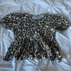 Leopardmönstrad T-shirt från Gina Tricot - Supersöt T-shirt från Gina Tricot med leopardmönster i beige och svart. Modellen har puffiga korta ärmar, volang nertill och knyt framtill. Perfekt för dig som vill sticka ut med ett djurmönstrat plagg och gillar trendiga detaljer.