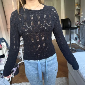 Svart stickad topp med öppen rygg - Snygg svart stickad topp med lång ärm och coolt hålmönster. Baksidan har en stor öppen rygg och knyt längst ner för extra detalj. Perfekt för dig som vill sticka ut med en unik och trendig look.