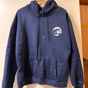 Mörkblå hoodie med måntryck - Snygg mörkblå hoodie med vit måne och stjärnor tryckt på bröstet. Tröjan har huva med snörning och en stor magficka framtill. Perfekt för dig som gillar rymdtema och vill ha en chill och bekväm stil.