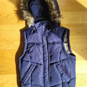 Mörklila dunväst med fuskpäls från H&M - Dunväst i mörklila från H&Mmed avtagbar huva och fuskpälsdetalj. Västen har två snedställda fickor med knappar och dragkedja framtill. Insidan är fodrad med rutigt tyg i beige och brunt. Perfekt för lager-på-lager under kyliga dagar 🍁