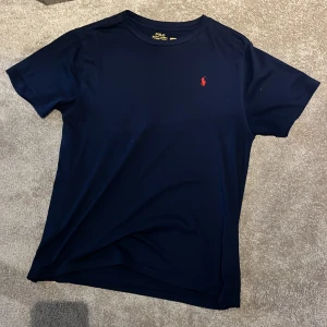 Mörkblå t-shirt från Polo Ralph Lauren - Snygg mörkblå t-shirt från Polo Ralph Lauren i storlek XL barn passar dig som brukar använda storlek S. Klassisk design med korta ärmar och röd broderad logga på bröstet. Tillverkad i mjuk bomull som är skön mot huden. Perfekt för dig som gillar stilrena och tidlösa plagg.