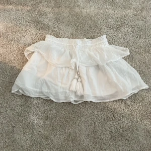 Vit volangkjol från Gina Tricot - Supersöt vit kortkjol från Gina Tricot med volangdetaljer och snörning i midjan med dekorativa tofsar. Kjolen är gjord i ett lätt och luftigt bomullsmaterial, perfekt för varma dagar. Elastisk midja för skön passform och flowy look. stl 140 men passar xs