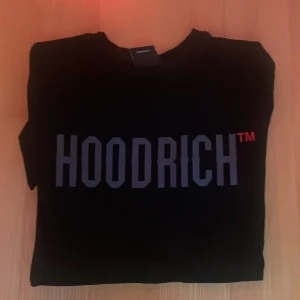 Svart HOODRICH t-shirt med tryck - Svart t-shirt från HOODRICH i storlek S med stor, grå logotyp  och  tryck på baksidan. T-shirten har rund hals . Materialet är mjuk bomull som känns skön mot huden. Perfekt för dig som gillar urban stil och tydliga prints. 