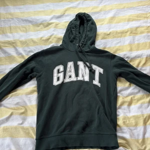 Mörkgrön hoodie från GANT med tryck - Snygg mörkgrön hoodie från GANT med stort vitt tryck över bröstet. Klassisk huva med snörning och långa ärmar. Perfekt för dig som gillar en avslappnad och sportig stil. Mjuk bomullskänsla och riktigt skön passform.
