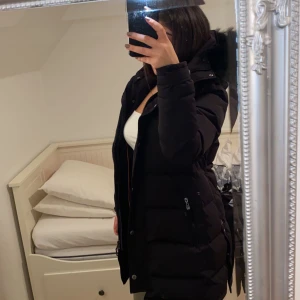 Svart lång dunjacka med huva Hollies  - Säljer en svart lång dunjacka från BoG med fluffig huva och dragkedja framtill. Jackan har en stilren look, är quiltad och har praktiska fickor med dragkedja. Perfekt för kalla dagar och har en snygg, mörk ton som passar till det mesta.