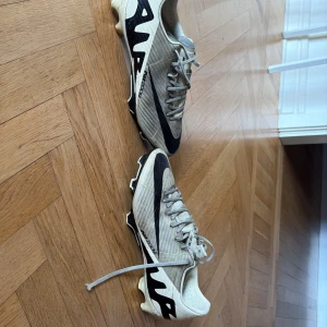 Nike fotbollsskor i beige och svart - Säljer ett par Nike fotbollsskor i beige med svarta detaljer och stor Nike-logga på sidan. Skorna har snörning och en platt sula med dobbar för bra grepp på planen. Ovandelen är tillverkad i syntetmaterial med ett diskret randigt mönster. Perfekt för dig som vill ha stil på fotbollsplanen.