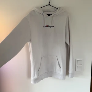 Vit hoodie Los Angeles tryck - Säljer en vit hoodie med coolt Los Angeles-tryck fram och en stor bild med eld och bil på ryggen. Tröjan har huva med snörning och en klassisk magficka. Perfekt för dig som gillar streetstyle och vill sticka ut med ett unikt motiv.