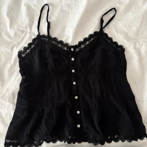Svart spetslinnetopp från Zara - Supersöt svart linnetopp från Zara med tunna axelband och fina spetsdetaljer längs kanterna. Toppen har knappar framtill och en lätt utsvängd passform. Perfekt att styla med jeans eller kjol för en trendig look. Inga defekter eller liknande! 