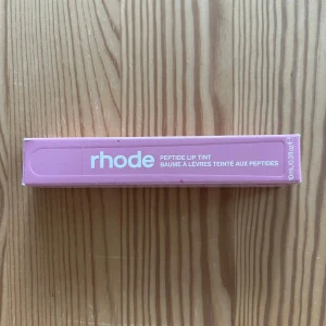 Rhode Peptide Lip Tint Ribbon - Rhode Peptide Lip Tint i färgen Ribbon, en rosa tonad läppbalsam med peptider. Kommer i en mjuk tub med vit text och rymmer 10 ml. Perfekt för dig som vill ha återfuktade läppar med en hint av färg och glans.