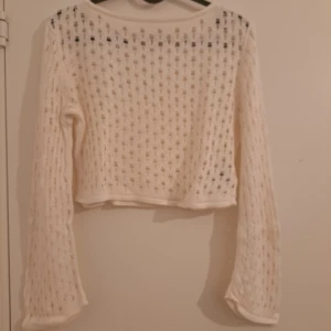 Vit stickad croppad topp från H&M - Vit stickad croppad topp från H&M med lång ärm och hålmönster över hela plagget. Toppen har en avslappnad passform och är perfekt för lager-på-lager-stil. Snygg att matcha med jeans eller kjol för en trendig look.