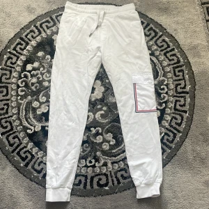  Moncler byxor - Snygga vita mjukisbyxor från Moncler i storlek S. Byxorna har resår och snörning i midjan, muddar vid bensluten och en cool ficka med knapp och röd-blå kant på benet. Perfekta för en chill och stilren look.