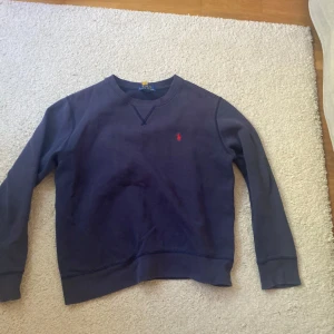 Mörkblå sweatshirt från Polo Ralph Lauren - Mörkblå sweatshirt från Polo Ralph Lauren med klassisk rund hals och broderad röd logga på bröstet. Tröjan har långa ärmar och ribbade muddar vid ärmslut och nederkant. Köpt på små hjärtan i Höllviken flr 1099 kr.