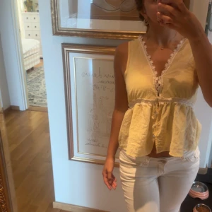 Gul ärmlös blus med spetsdetaljer - Söt gul blus med v-ringning och vita spetsdetaljer längs halsringningen och midjan. Modellen är ärmlös och har en lätt volang nedtill som ger en luftig känsla. Perfekt för varma dagar och enkel att styla med jeans eller kjol. Den ör mer öppen vid brösten men har en säkerhets nål