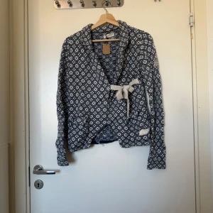 Blåvit mönstrad stickad kofta med knyt - Säljer en blåvit stickad kofta från Odd Molly med snyggt geometriskt mönster och omlottknytning i midjan. Koftan har långa ärmar, mjukt material och coola detaljer som ribbad kant och fickor framtill. Perfekt för dig som gillar unika plagg med lite extra stil. Storlek 2 som enligt deras storleksguide motsvarar en storlek M.  Fint skick, men tyvärr finns lösa trådar på insidan och små tecken på användning. 