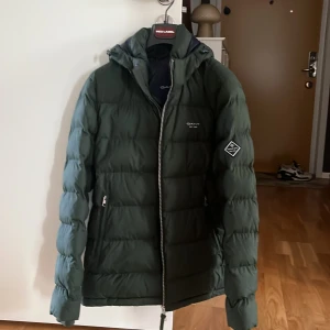 Mörkgrön pufferjacka från GANT M - Säljer en mörkgrön pufferjacka från GANT i storlek medium. Jackan har huva med dragsko, två sidofickor med dragkedja och GANT-logga på bröstet och ärmen. Perfekt för kalla dagar med sin quiltade design och snygga look. Storleken kan även passa en liten L. Original pris: 3000. Köpt för ett år sen men knappt använd på grund av en ”varm vinter”. Pris kan diskuteras vid snabb affär. 